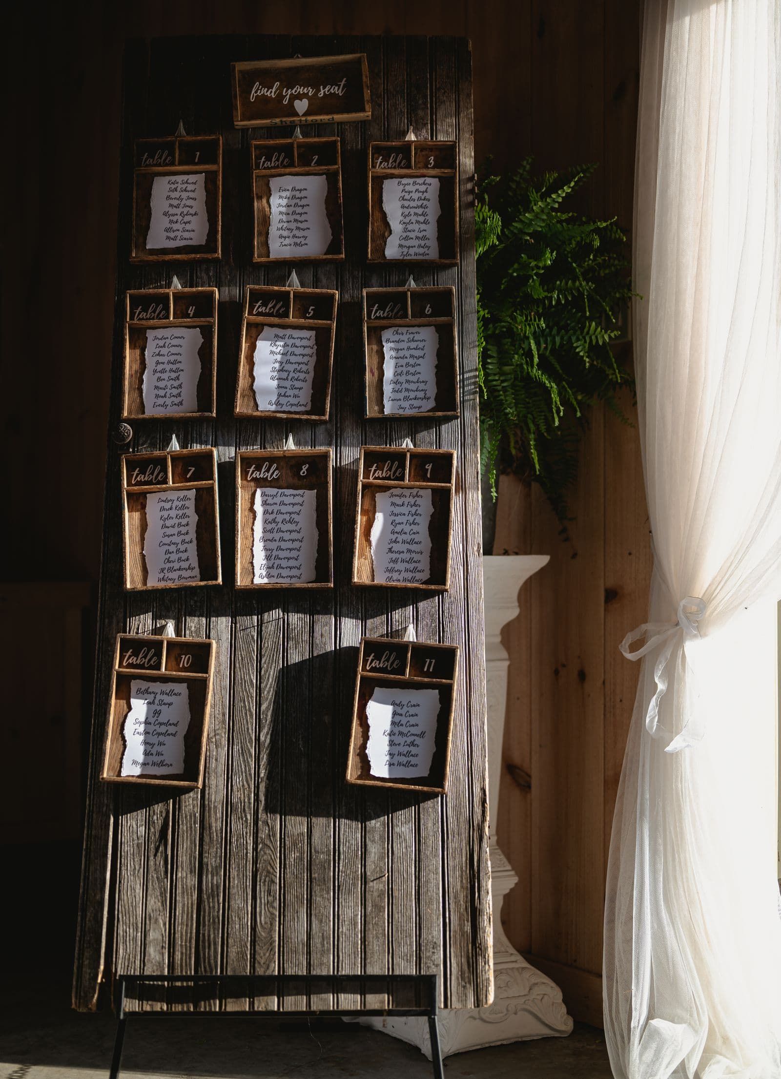 Rustic wooden wedding seating chart displayed beside a potted plant in the sunlight